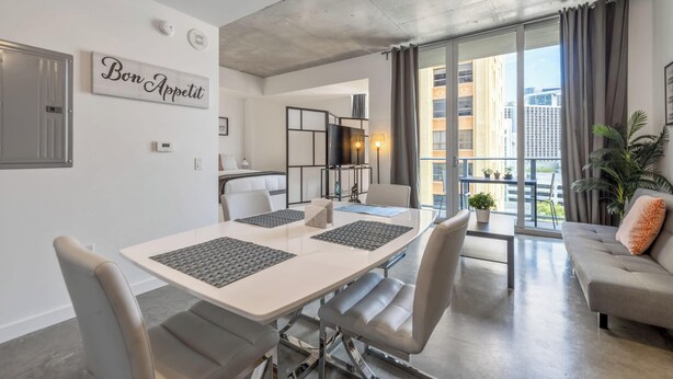 A modern living room with a dining area, a sofa, a large window with curtains, and a balcony with a view of buildings.