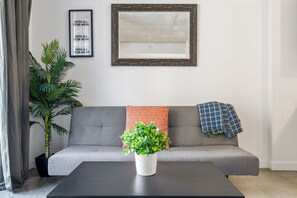 Ein Wohnzimmer mit grauem Sofa, einem weißen Blumentopf, einem schwarzen Couchtisch, einem gerahmten Zitat an der Wand und einem großen Spiegel.