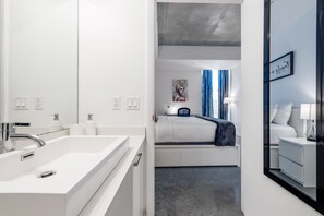 Quarto de hotel moderno com um espelho grande, uma pia branca, uma cama com manta azul e uma mesa de cabeceira com abajur.