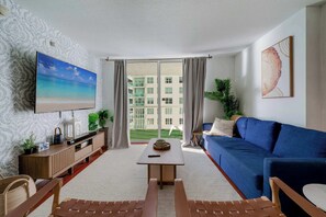 A living room with a TV, sofa, coffee table, and a view of a building through large windows. - Brickell Bay View- Unbeatable Location With Parking (Miami)