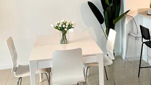 Et spiseområde med et hvidt bord, fire stole, en vase med blomster, et vægur og en stor plante.