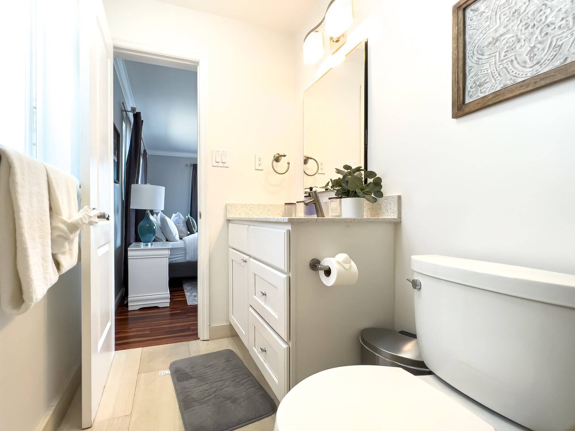 A compact bathroom with a toilet, sink, and mirror, connected to a bedroom with a bed and a nightstand.