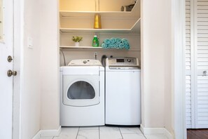 A laundry room with two washing machines, a shelf with a green bottle and a roll of blue towels, and a white door.