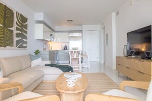 A modern living room with a sofa, coffee table, and TV. There is a kitchen in the background with white cabinets and a dining area. - Brickell House Dreams - Luxury Stay and Amenities (Miami)
