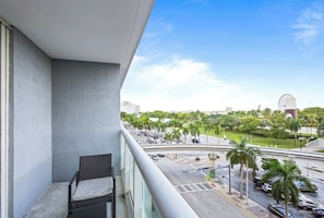A balcony with a wicker chair, a view of a city street with palm trees, and a large Ferris wheel in the distance. - Biscayne Bay Views-pool, Hot Tub and Amenities (Miami)