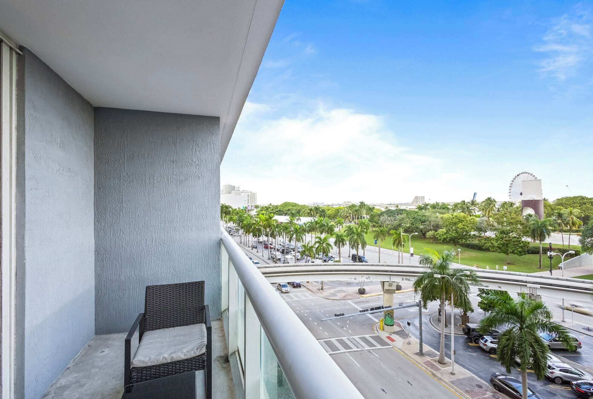 Un balcone con una sedia in vimini, vista su una strada cittadina con palme e una grande ruota panoramica in lontananza.