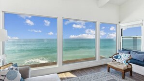 Ein Zimmer mit großen Fenstern und Meerblick, eine hölzerne Bank sowie ein Sofa mit gemusterten Kissen.