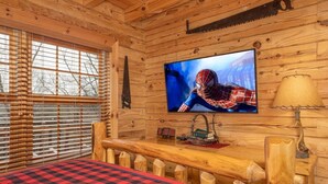 A log cabin room with a television, a bed with a checkered blanket, a wooden bench, and a window with blinds. - 492a) Mountain View 4BR With Hot Tub! (Sevierville)