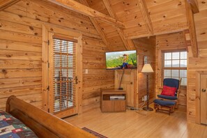 Ein Zimmer einer Holzhütte mit einem Bett, einem an der Wand befestigten Fernseher, einem Sessel und einer Lampe.