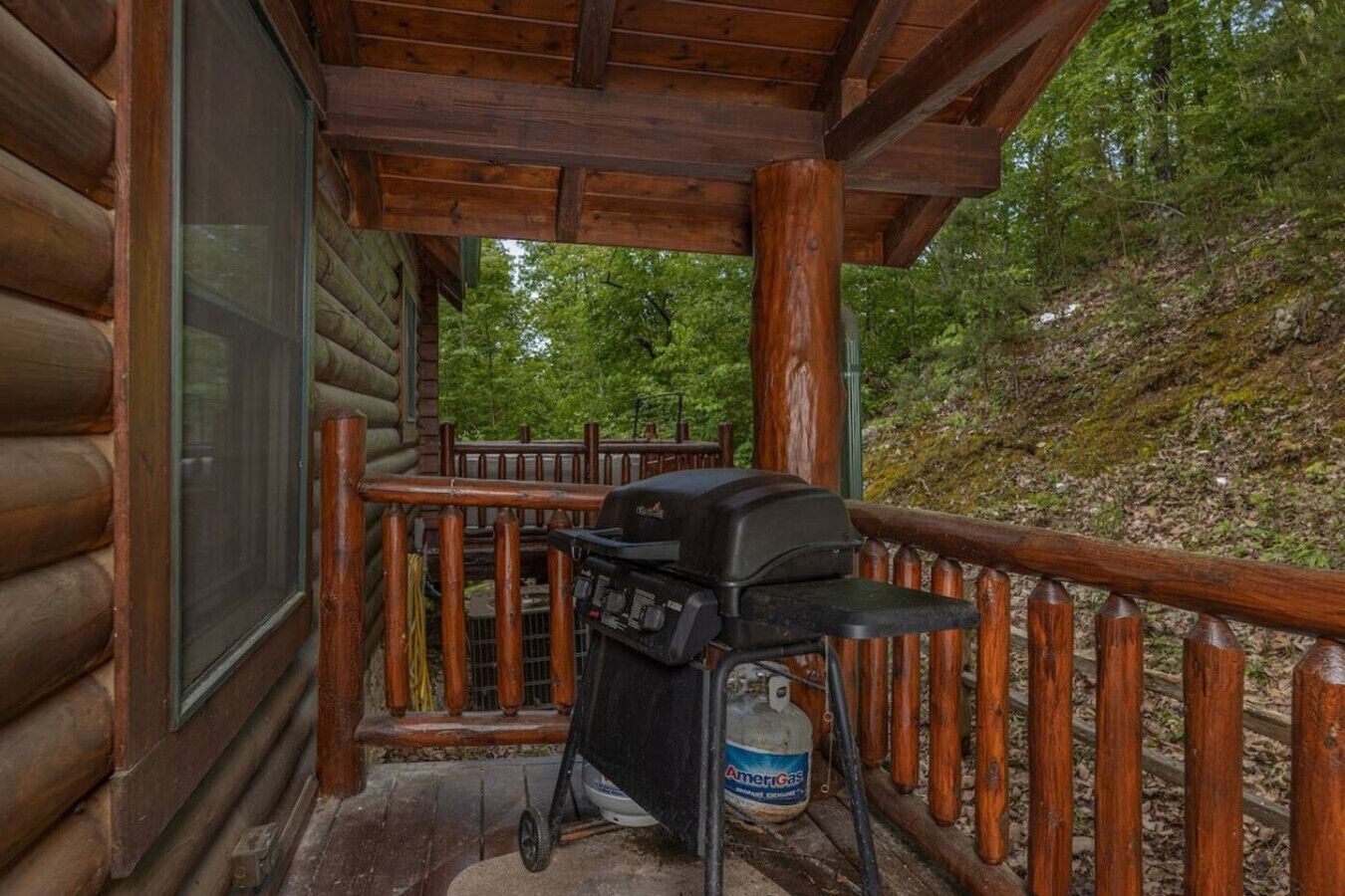 A wooden porch with a grill and propane tank, surrounded by a wooden railing and a view of a forested hillside.