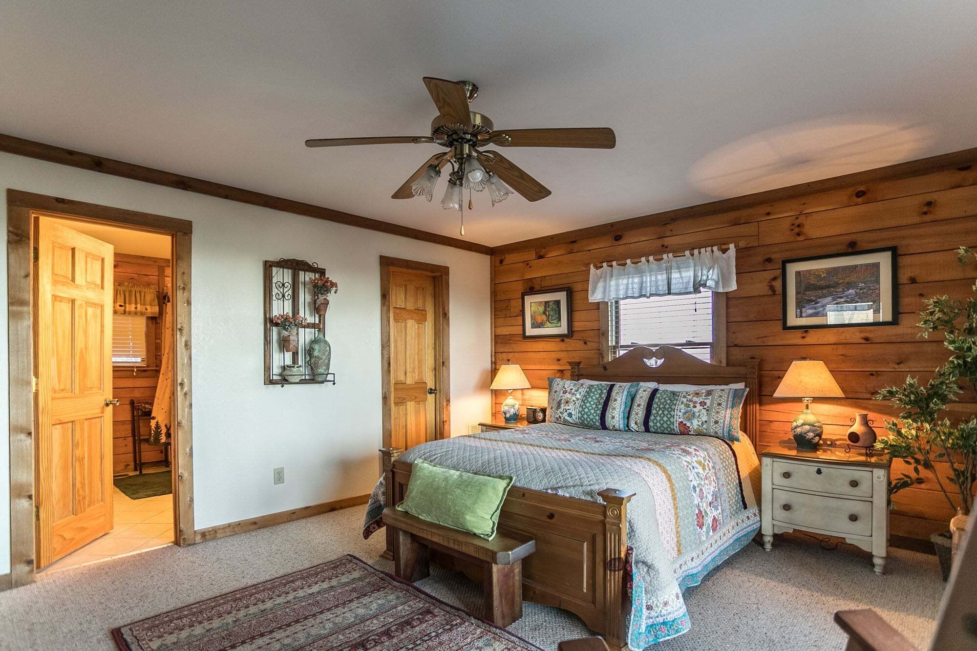 A bedroom with a wooden ceiling fan, a bed with a quilt, a nightstand with lamps, and a window with curtains.