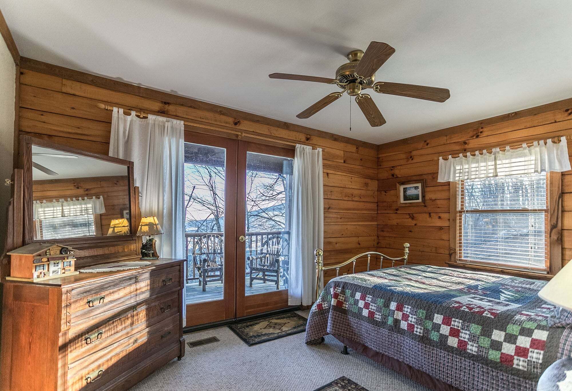 A wooden cabin bedroom with a bed, dresser, ceiling fan, and large windows.