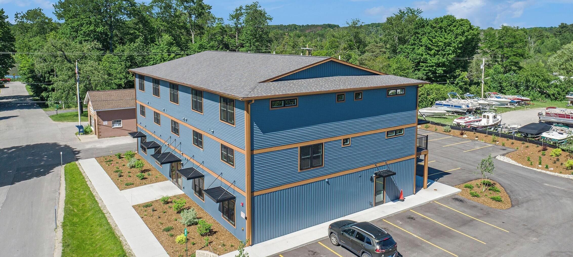 A multi-story building with a blue exterior, a parking lot, and boats in the background.