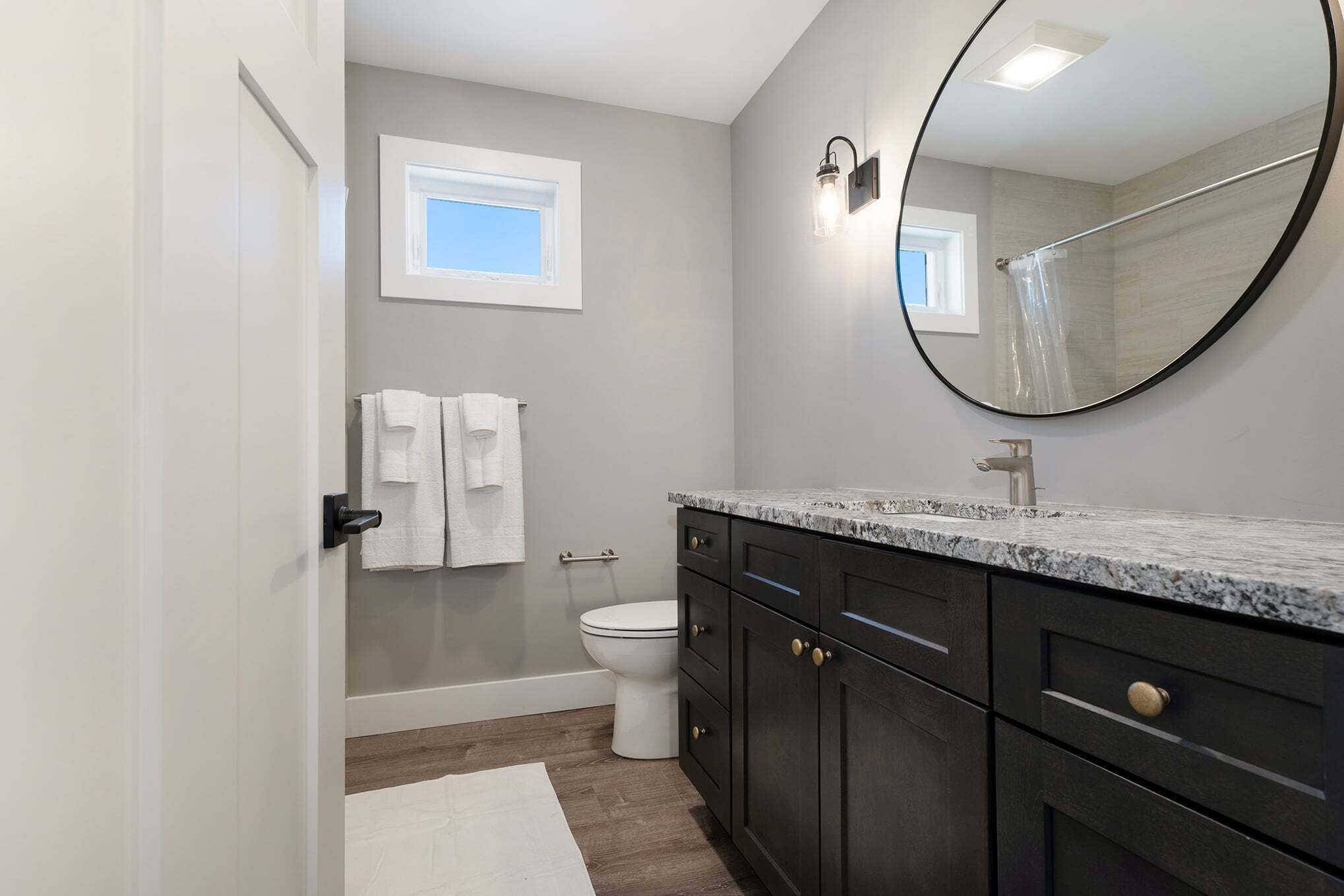 A bathroom with a toilet, a sink with dark cabinets, a mirror, and a window.