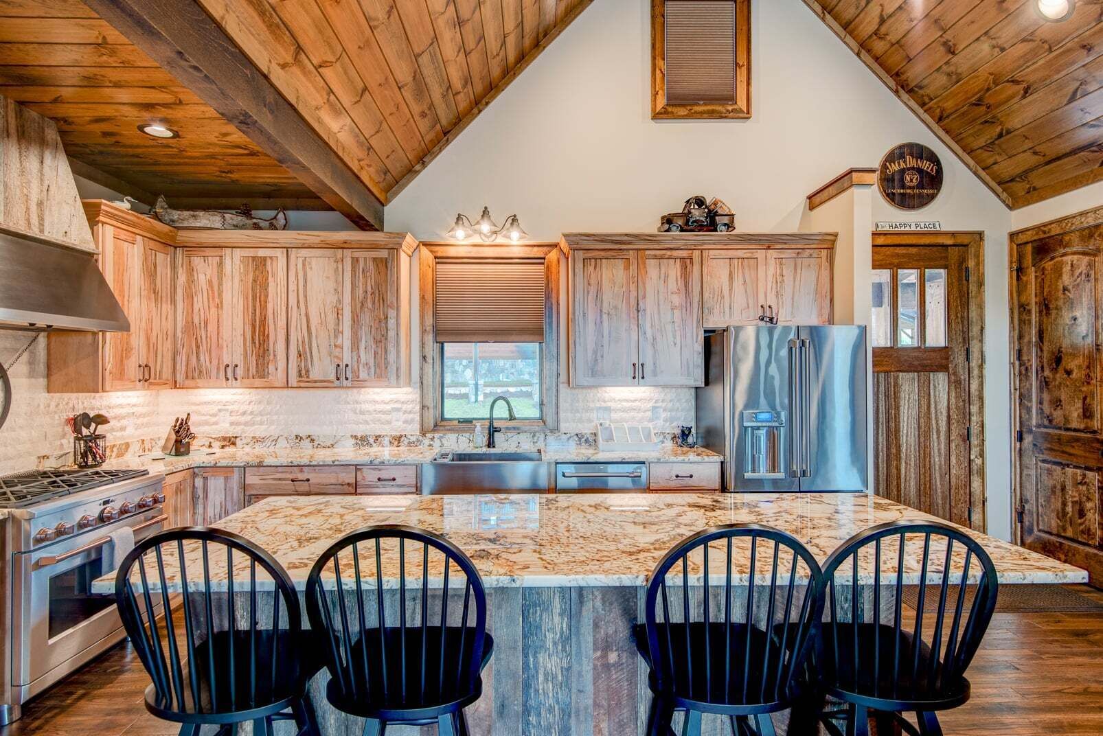 A kitchen with wooden cabinets, a stainless steel refrigerator, a large island with bar stools, and a high ceiling with exposed beams.
