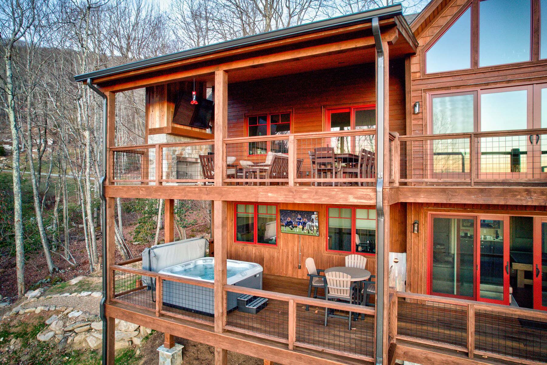 A multi-level wooden cabin with a hot tub, outdoor seating, and a large window displaying a sports event.