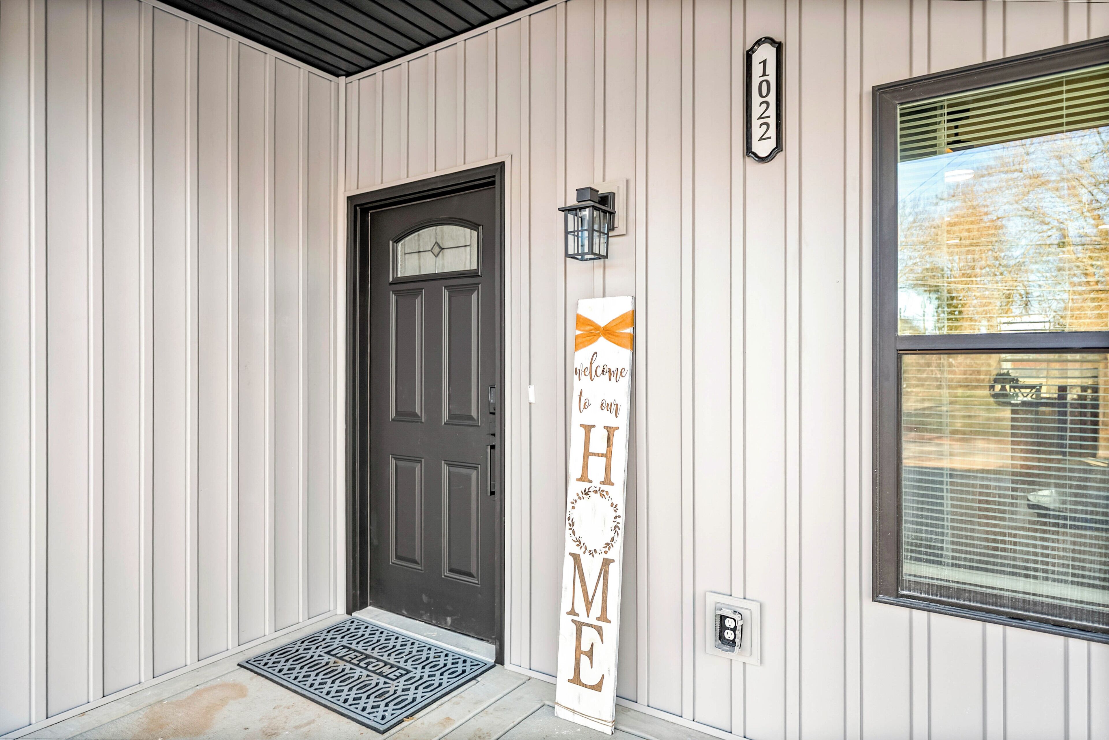A house entrance with a dark door, a welcome sign, a mailbox, and a window with blinds.