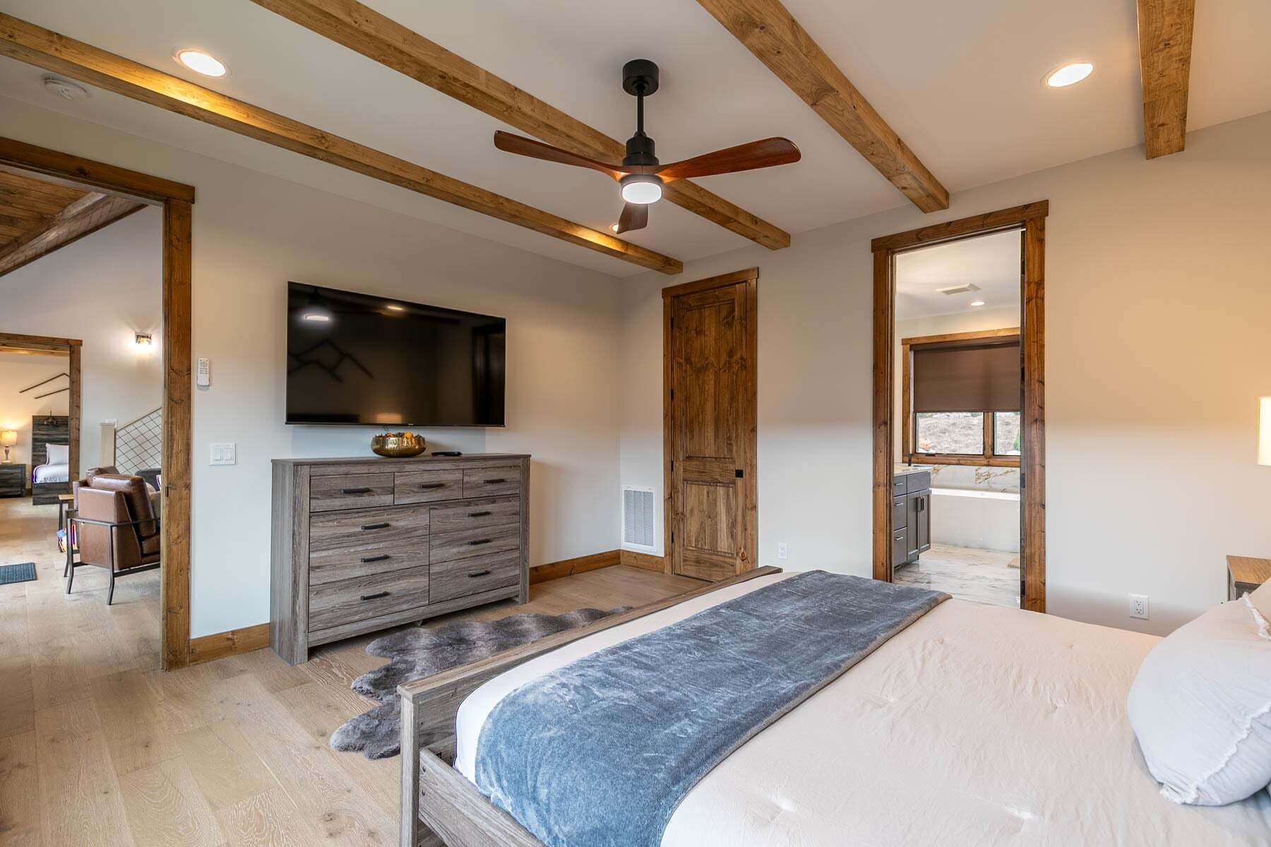 A bedroom with a bed, a dresser, a ceiling fan, a TV, and a door leading to another room.