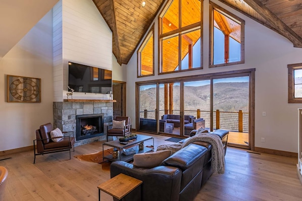 A spacious living room with a fireplace, leather furniture, and large windows offering a view of the outdoors. - Mountain Time Lodge at Eagles Nest (Banner Elk)