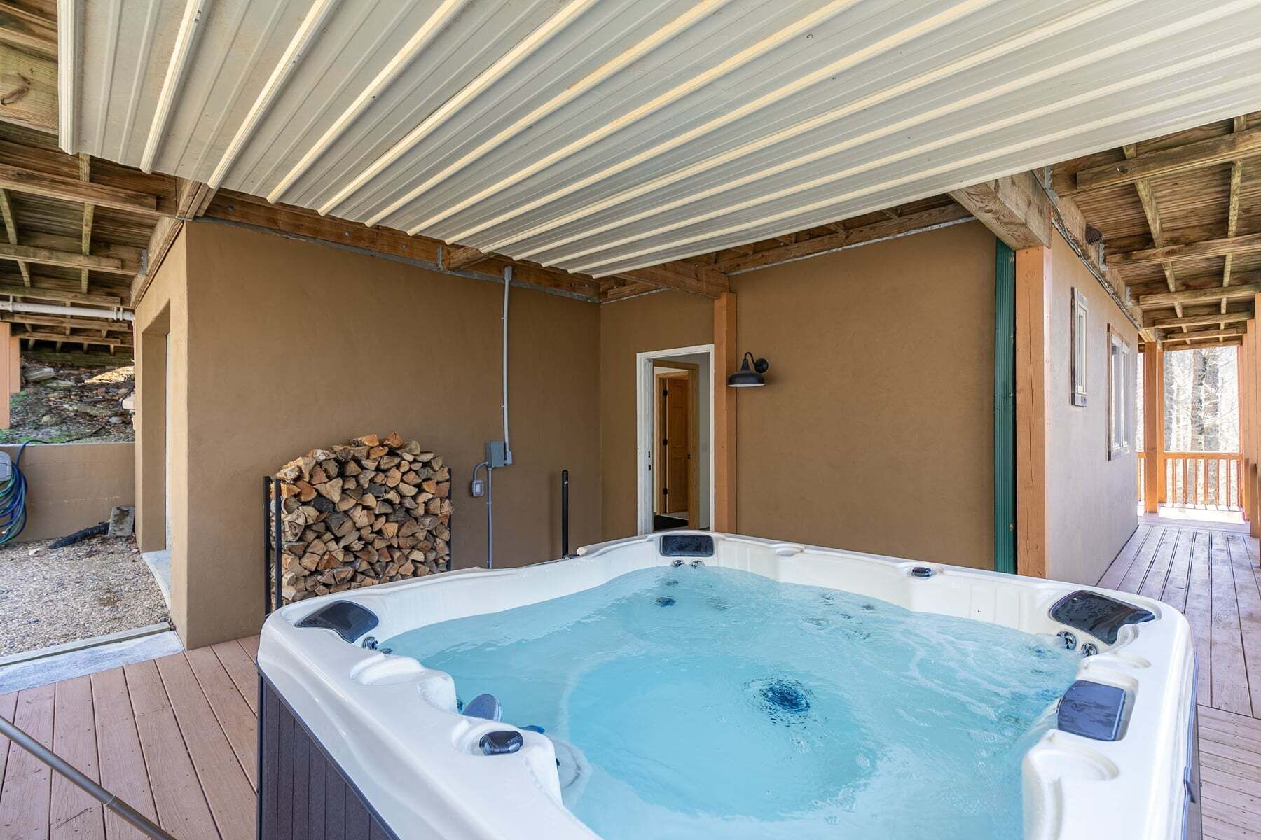 Un bain à remous dans un espace extérieur abrité, doté d’une terrasse en bois, d’une pile de bois de chauffage et d’une porte donnant sur une autre pièce.