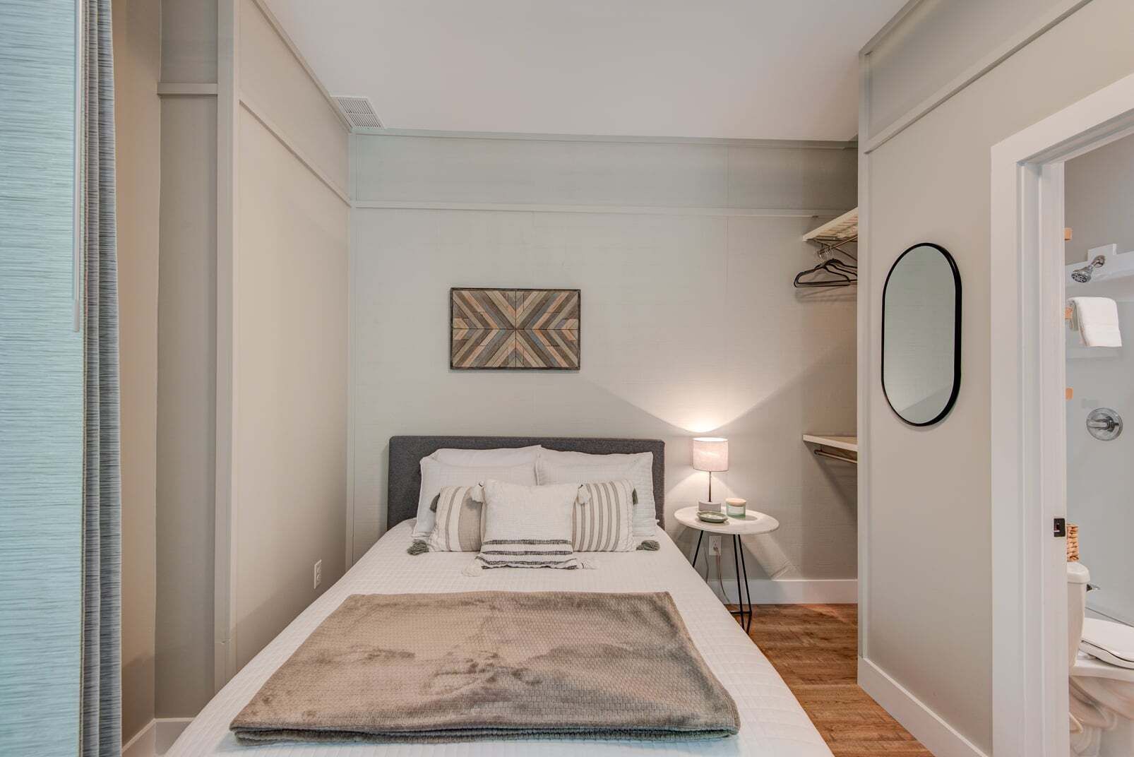 A narrow hotel room with a bed, a bedside table with a lamp, a mirror, and a wall-mounted artwork.