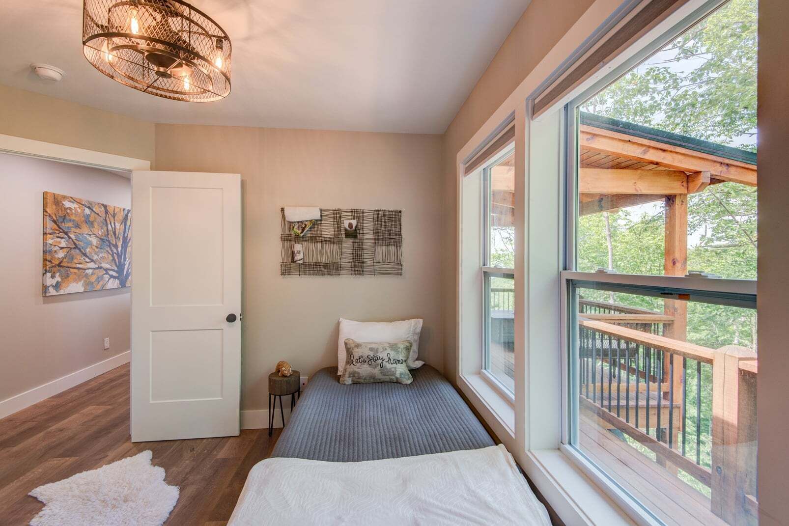 A bedroom with a bed, a white door, a wall-mounted artwork, a ceiling light fixture, and a large window with a view of a deck.