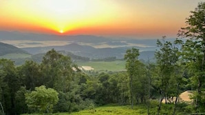 A circular outdoor seating area with a fire pit, surrounded by lush greenery and a view of distant hills during sunset. - Amongst the Clouds at Chappell Farms (Banner Elk)