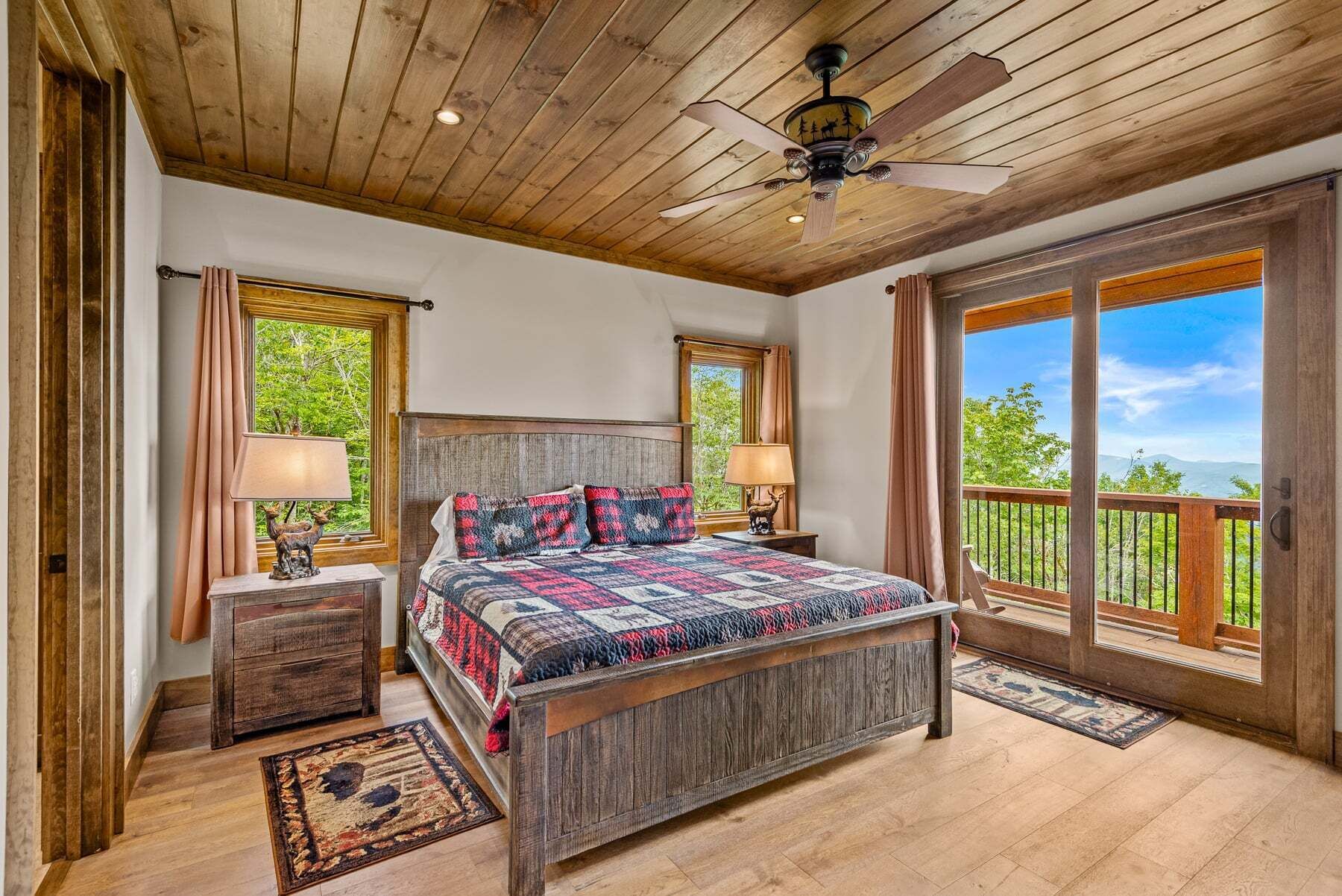 Une chambre avec un plafond en bois, un lit recouvert d’une couverture à motifs, deux tables de chevet avec des lampes et une vue dégagée sur l’extérieur grâce à une grande fenêtre.