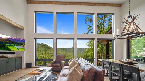 A spacious living room with a large window, leather sofa, and a dining area with a wooden table. - Sky's the Limit at Blue Ridge Mountain Club (Boone)