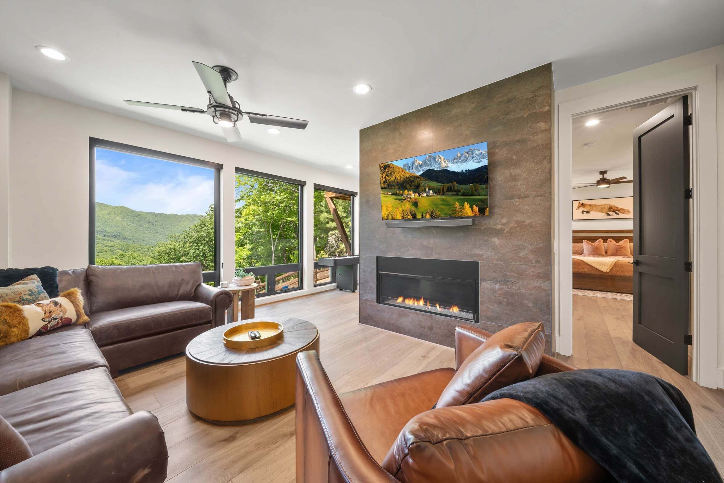A modern living room with a fireplace, leather sofas, a coffee table, and large windows offering a view of greenery.
