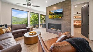 A modern living room with a fireplace, leather sofas, a coffee table, and large windows offering a view of greenery. - Sky's the Limit at Blue Ridge Mountain Club (Boone)