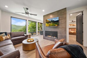 Ein modernes Wohnzimmer mit Kamin, Ledersofas, einem Couchtisch und großen Fenstern mit Blick auf viel Grün.