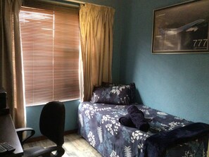 Ein Schlafzimmer mit einem Einzelbett, einem Sessel, einem Schreibtisch und einem Fenster mit Jalousien.