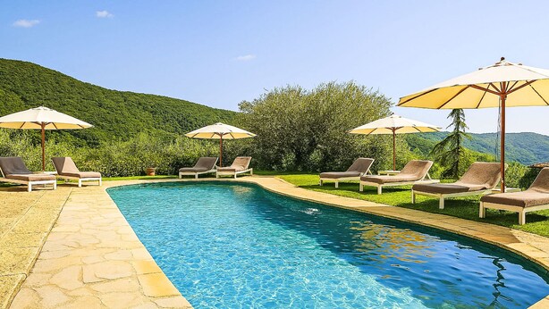 Ein Schwimmbecken mit Liegestühlen und Sonnenschirmen, umgeben von Grünflächen und Hügeln.