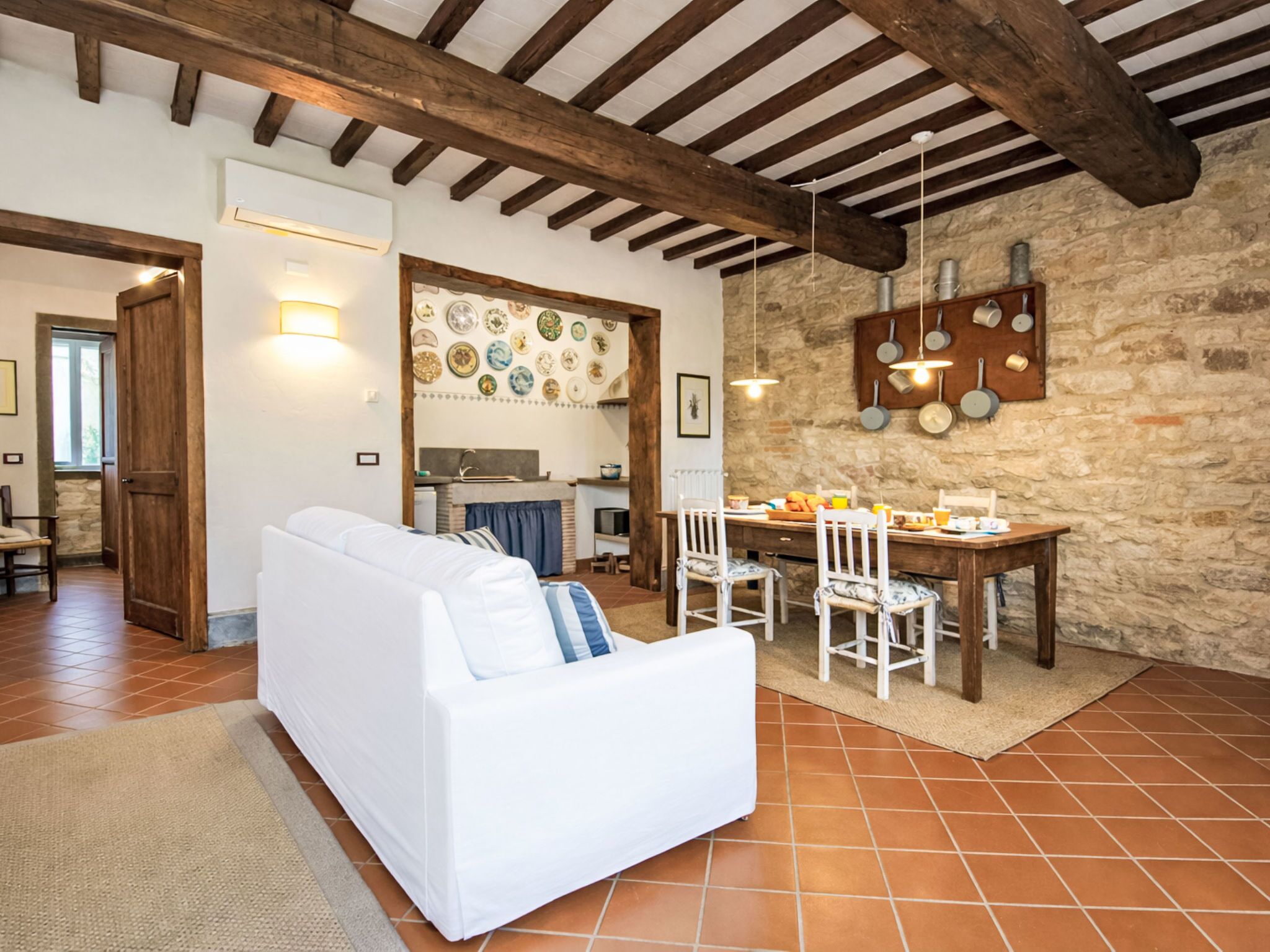 Una sala de estar acogedora con un sofá blanco, una mesa de comedor con sillas, una zona de cocina con paredes de piedra y vigas de madera a la vista en el techo.