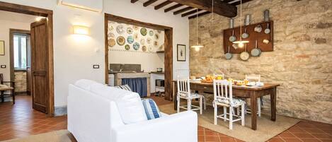 Una sala de estar acogedora con un sofá blanco, una mesa de comedor con sillas, una zona de cocina con paredes de piedra y vigas de madera a la vista en el techo.