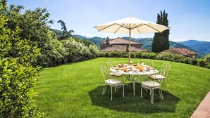 Een verzorgd gazon met een ronde tafel en stoelen, een grote parasol en een bergachtig landschap op de achtergrond.