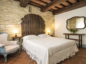 A bedroom with a stone wall, wooden ceiling beams, a bed with white bedding, a chair, a bedside table with a lamp, and a mirror on the wall. - Le Mangiatoie by Interhome (Rufina)