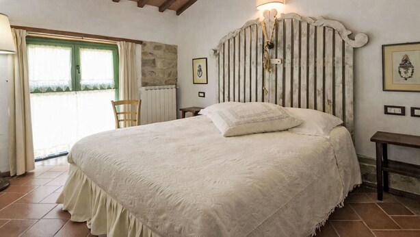 A bedroom with a large bed, wooden ceiling beams, a stone wall, a window with curtains, and a small bedside table. - Il Pastore by Interhome (Rufina)