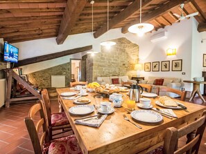 A dining area with a wooden table set for breakfast, stone walls, and a television in the background. - Il Pastore by Interhome (Rufina)