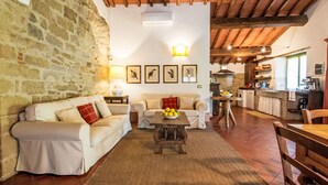 A cozy living room with a stone wall, wooden ceiling beams, a fireplace, and a kitchen in the background. - Il Pastore by Interhome (Rufina)