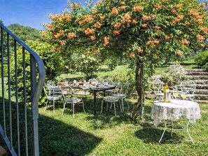 An outdoor dining area with tables and chairs, surrounded by lush greenery and a tree with orange flowers. - La Noce by Interhome (Rufina)