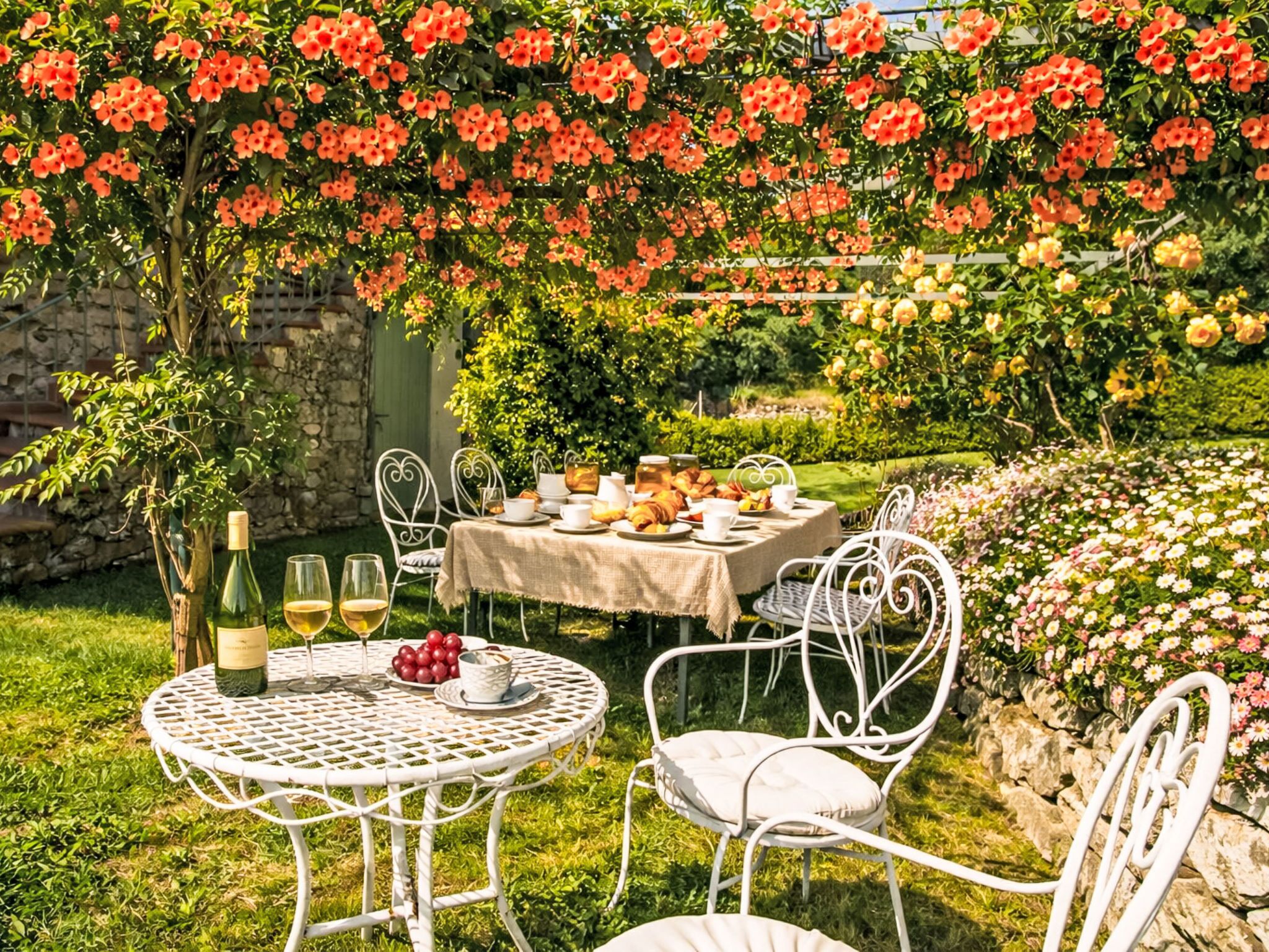 An outdoor dining area with round tables, white chairs, and a variety of flowers in the background.
