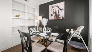 Un coin repas avec une table en verre, des chaises noires, une étagère blanche ornée d’objets décoratifs, un vase blanc et un tableau représentant un cheval accroché au mur.