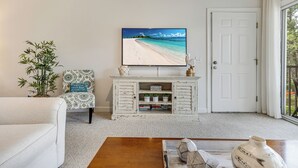 A living room with a television displaying a beach scene, a white couch, a patterned armchair, a wooden coffee table, and a white door. - Condo in Sea Trail, GREAT GOLF view, Restaurants on site, pool at the complex.  (Sunset Beach)
