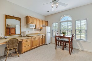 Una cocina con armarios de madera, microondas, refrigerador, mesa de comedor con sillas, ventilador de techo y un amplio ventanal con persianas.