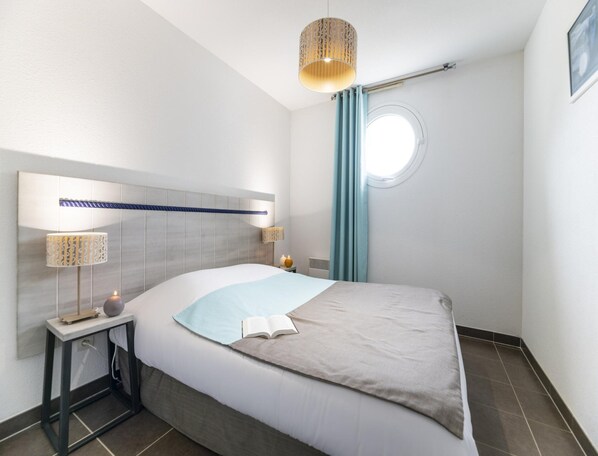 A modern bedroom with a bed, bedside lamps, a round window, and a headboard. - Les Carrelets 3 Bedroom Split-level APT (Saint-Palais-sur-Mer)