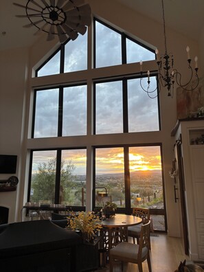 A room with large windows, a chandelier, and a dining area with chairs and a table.