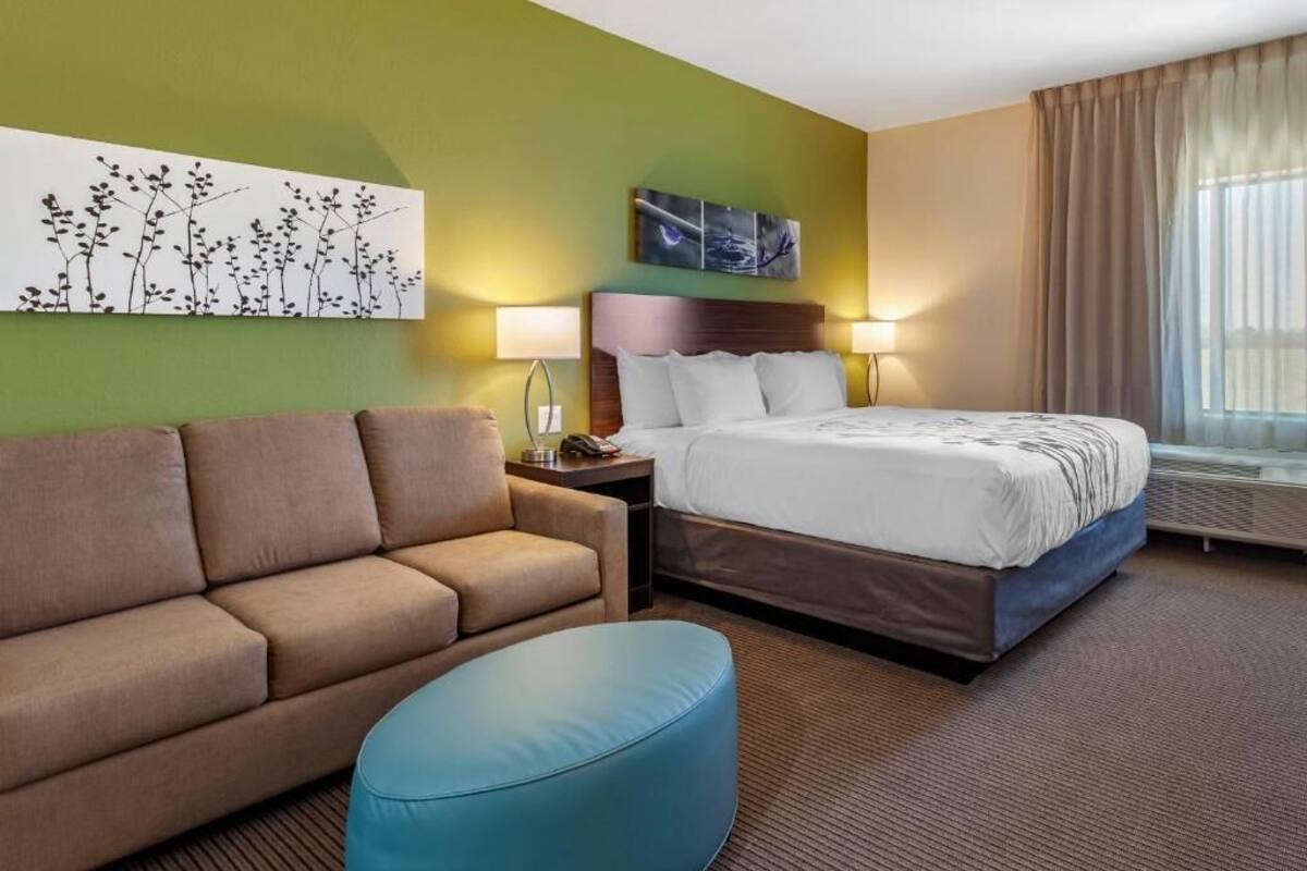Una habitación de hotel con una cama, un sofá, un puf azul, una mesita de noche, dos lámparas de pared y una ventana con cortinas.