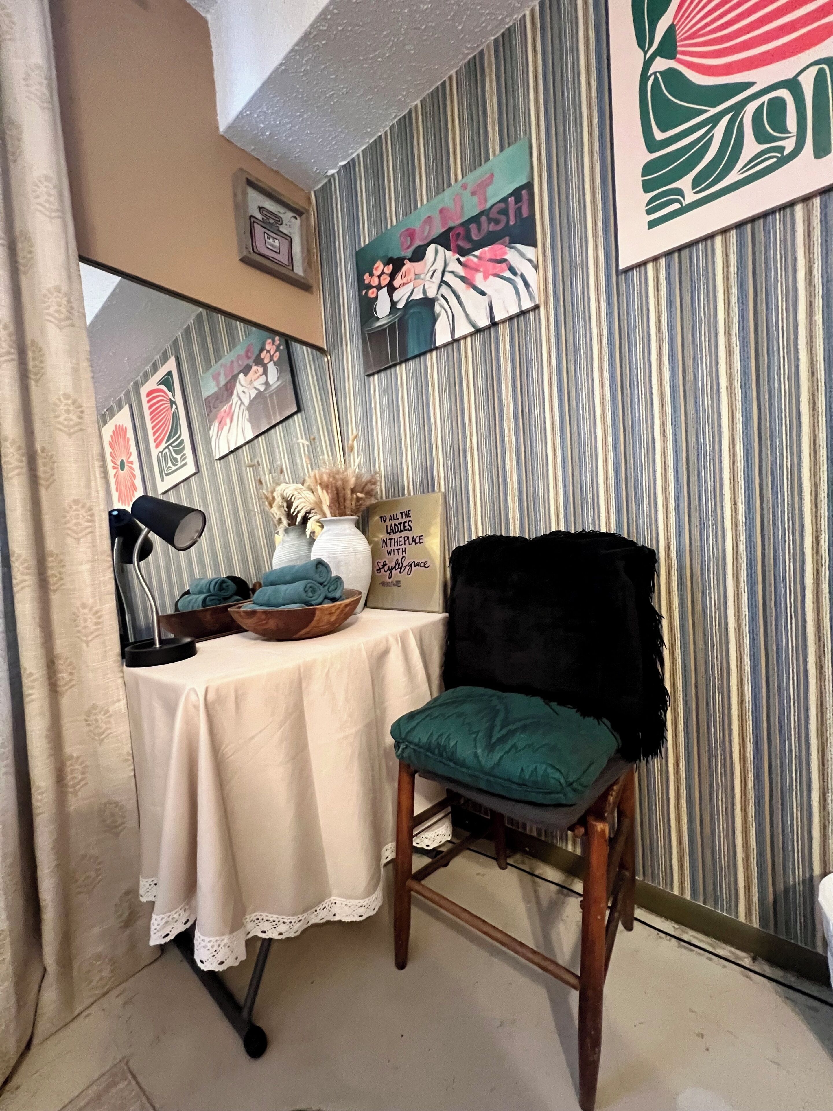 Ein Zimmer mit gestreifter Tapete, ein Tisch mit Schüssel und aufgerollten Handtüchern, ein Stuhl mit grüner Sitzkissen und eine Wand mit dem Schriftzug 'Don't Rush'.